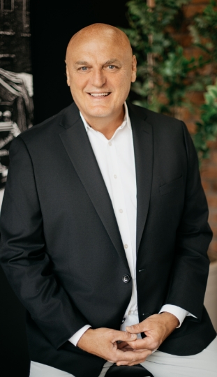 A smiling, bald man in a dark blazer and white shirt sits in front of a leafy plant and a dark background, looking confidently at the camera.