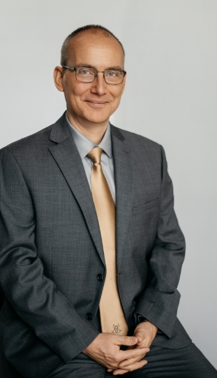 A middle-aged man with glasses, short hair, and a slight smile is wearing a gray suit, light gray shirt, and beige tie, seated against a plain light background.