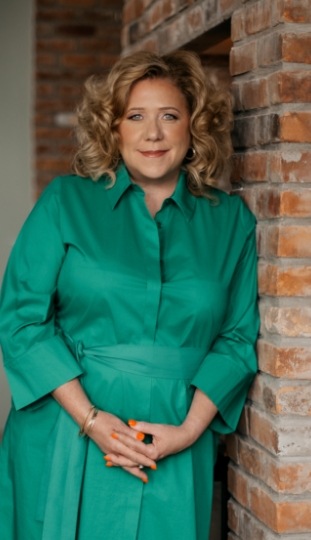 A woman with curly blonde hair, wearing a green dress, stands and smiles while leaning against a brick wall. She has orange nail polish and her hands are clasped in front of her.