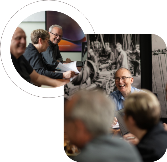 A group of people in a meeting, smiling and conversing around a table. The focus is on a man in glasses, appearing engaged and cheerful, with blurred colleagues in the foreground.