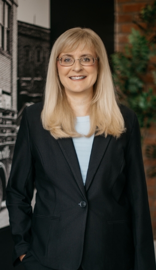 A woman with long blonde hair and glasses is smiling while wearing a dark suit jacket and light blue shirt, standing indoors in front of a black-and-white photo and green plants.