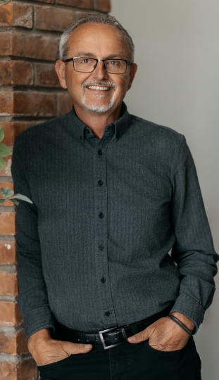 A smiling older man with gray hair, glasses, and a short beard stands by a brick wall, wearing a dark button-up shirt and black pants with his hands in his pockets.