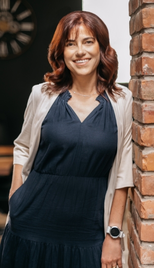 A woman with shoulder-length auburn hair, wearing a navy dress and light blazer, stands smiling and leaning against a brick wall. She has one hand in her pocket and wears a smartwatch on her wrist.
